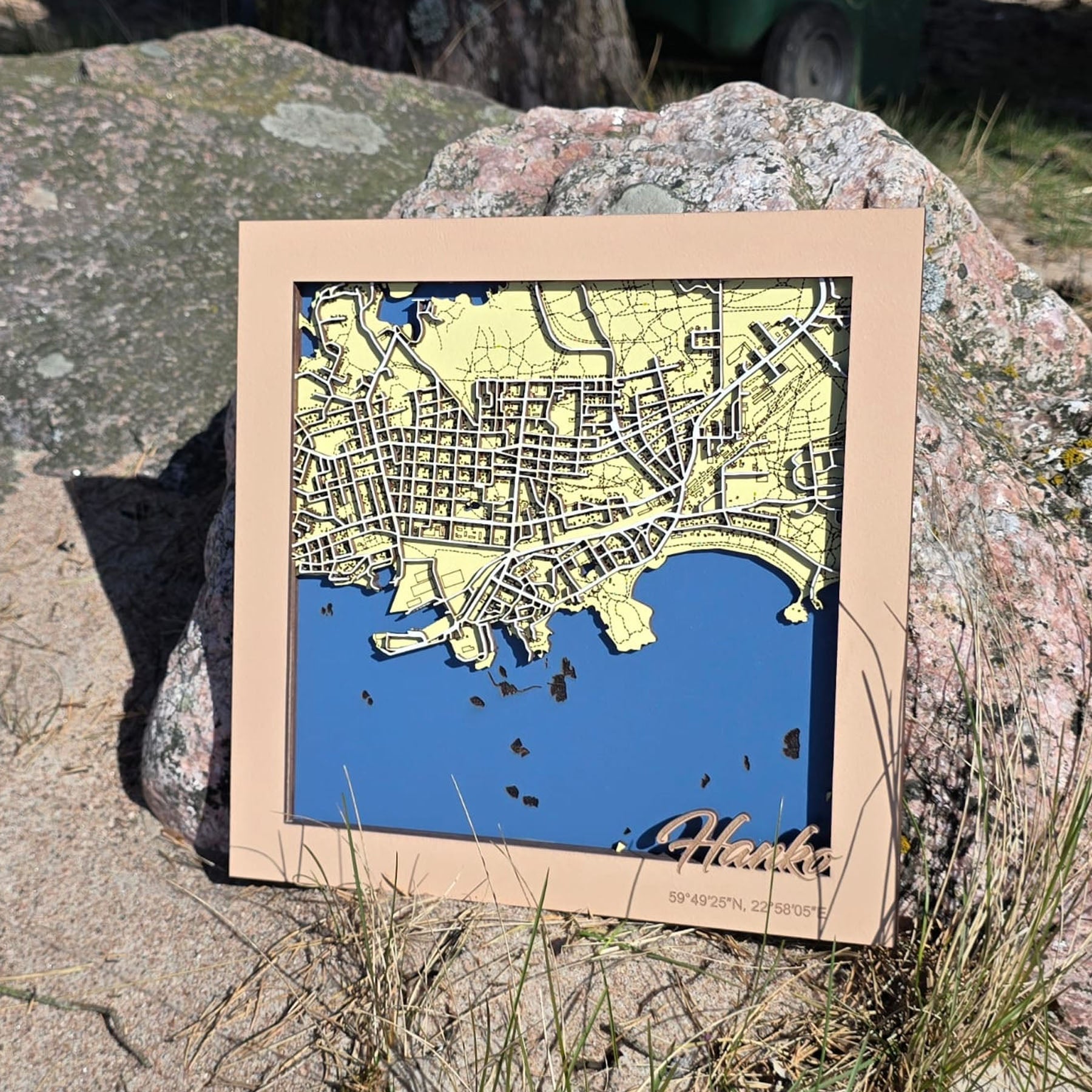 Handgjord träkarta över Hangö/Hanko med detaljerad stadsbild, blått hav och dekorativ träram – fotograferad i skärgårdsmiljö. Käsintehty puukartta Hangosta, yksityiskohtainen kaupunkikuva, sininen meri ja koristeellinen puukehys – kuvattuna saaristomaisemassa. Handmade wooden map of Hanko/Hangö featuring a detailed city layout, blue sea and decorative wooden frame, photographed in an archipelago setting.