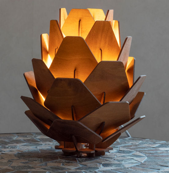 A table lamp with a pine cone shape, made from birch veneer, with light shining through, casting a warm glow. The lamp is placed on a textured surface.