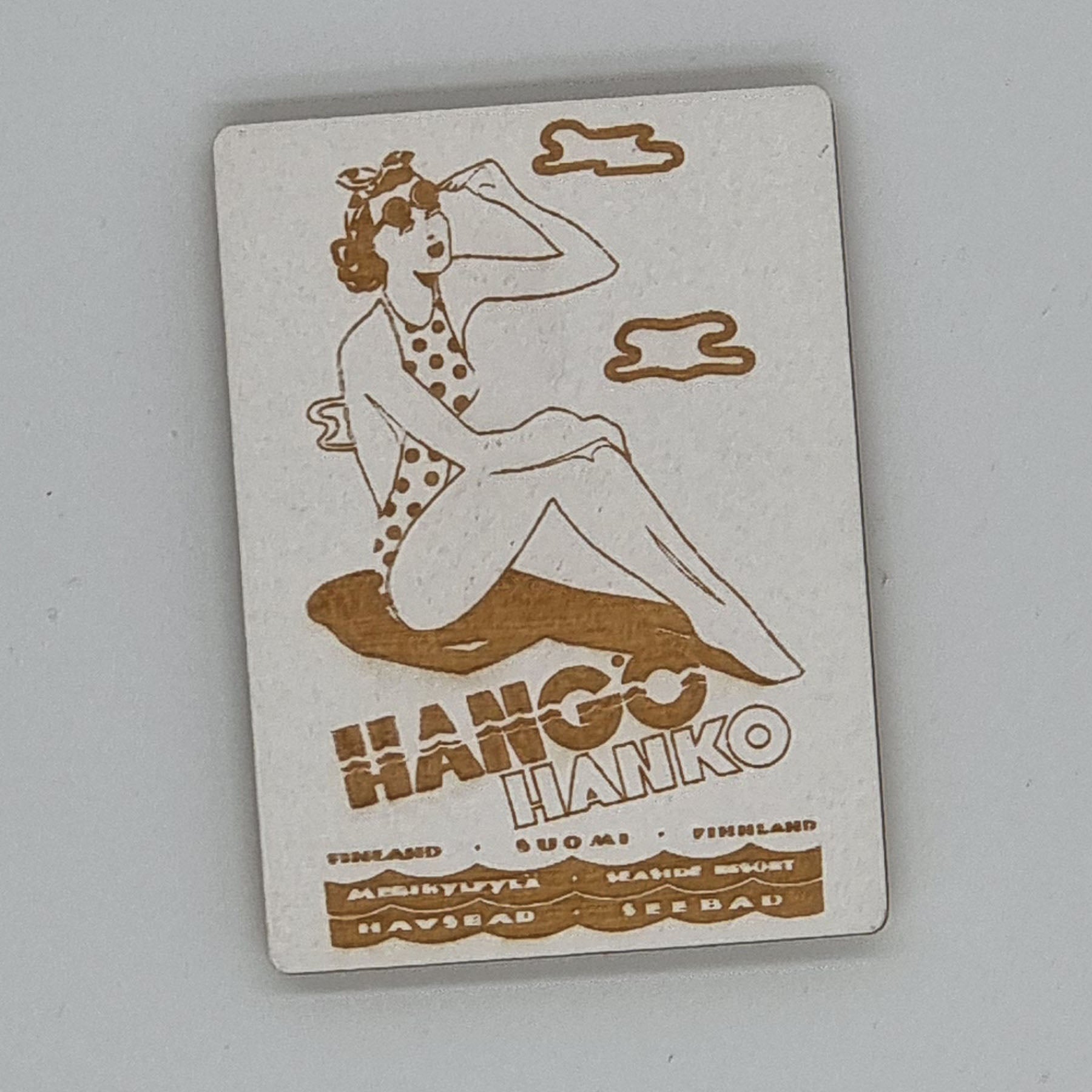 A magnet featuring a cartoon-style woman with a hat sitting on a bench, surrounded by text that includes 'HANGO HANKO' and other Finnish words.