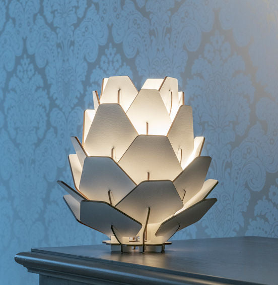 A table lamp with a design resembling a pine cone, made from birch veneer, with light shining through, casting a warm glow.