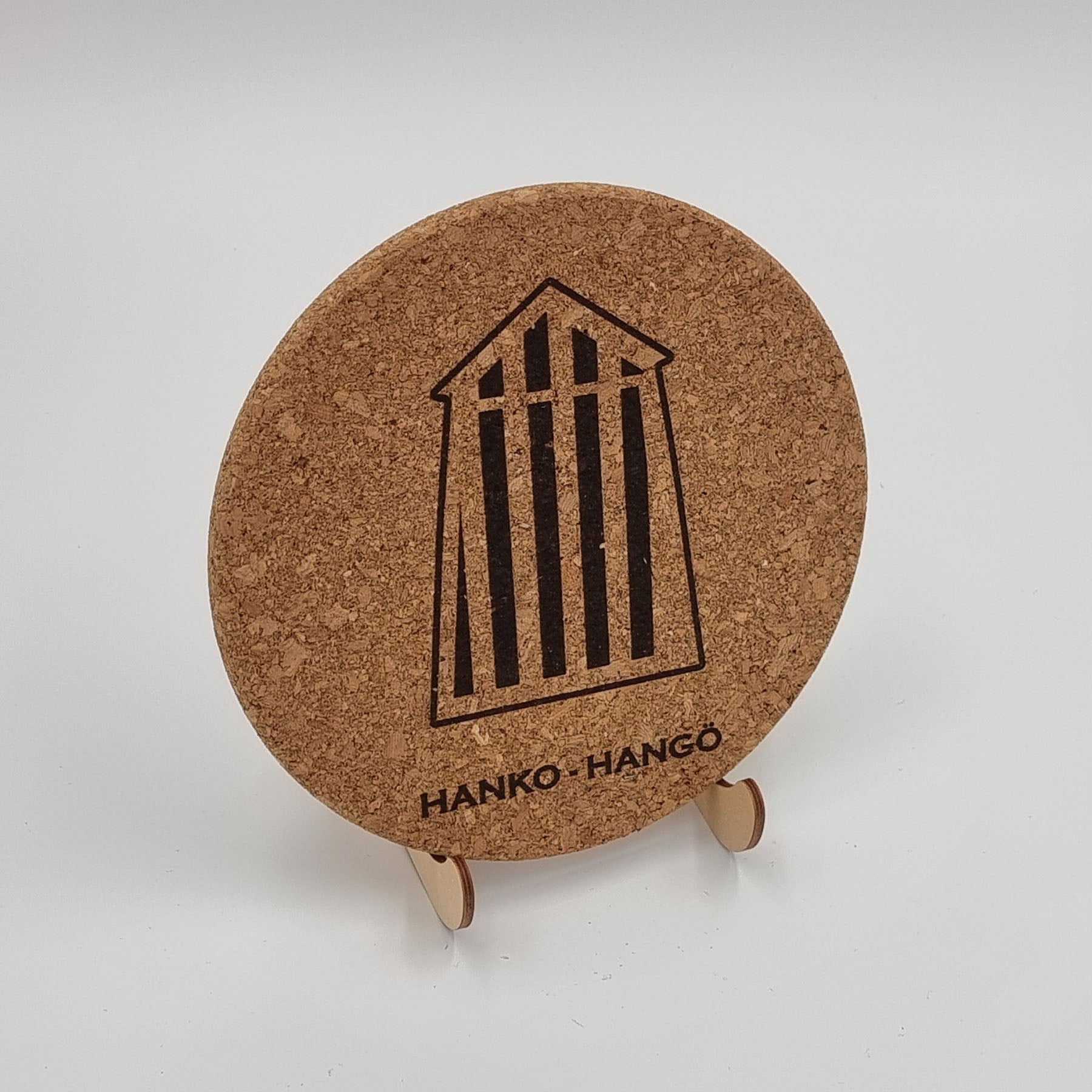 A round natural cork trivet with a engraved design of a bathing cabin and the text 'HANKO HANGO'.